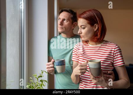 Serious concern young family couple standing together by window at home drinking morning coffee Stock Photo