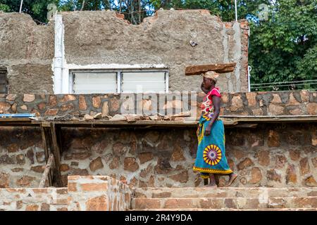 A construction worker carries a plank on her head down stone steps, wiping her brow with her free hand. Men and women are working together to build a stone house on the grounds of Kumbali Country Lodge near Lilongwe, Malawi. Stock Photo