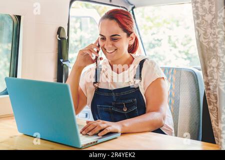 Woman working on her laptop and having a phone call in her handmade camper van while traveling. Van road trip holiday and outdoor summer adventure. No Stock Photo