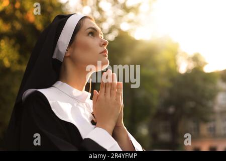 Young nun with hands clasped together praying outdoors on sunny day, space for text Stock Photo