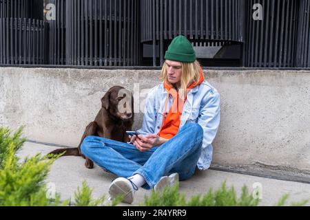 Sad young lonely man sitting on city street with dog using smartphone, having life troubles Stock Photo