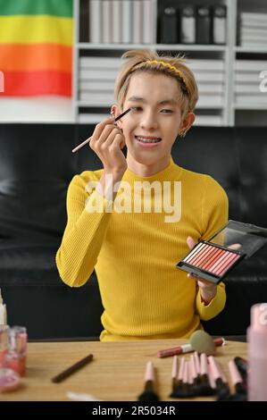 A portrait of an attractive young Asian gay beauty blogger or influencer is applying eyeshadow to his eyelid while recording his makeup tutorial video Stock Photo