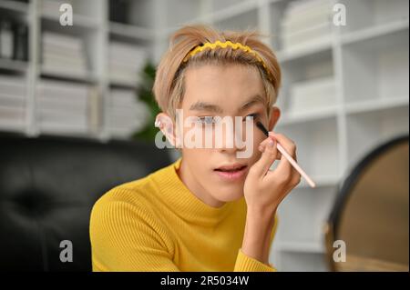 An attractive young Asian gay man is looking at the mirror and applying eyeshadow to his eyelid while doing his makeup at home. Stock Photo