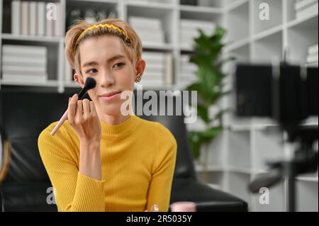 A happy and attractive young Asian gay beauty blogger or influencer is applying a brush to his cheeks and recording his everyday makeup look tutorial Stock Photo