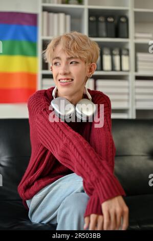 A portrait of an attractive and happy young Asian gay man in casual clothes sits on a sofa in his modern living room. LGBTQ+ lifestyle concept Stock Photo