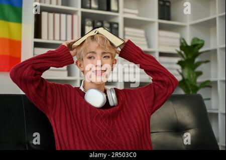 Portrait of an attractive and happy young Asian gay man in casual clothes sits on the sofa in the living room with a book. Stock Photo