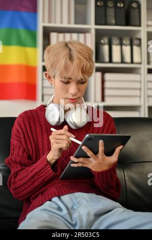 A portrait of an attractive young Asian gay man in casual clothes focuses on something on his tablet screen while sitting on the sofa in the living ro Stock Photo