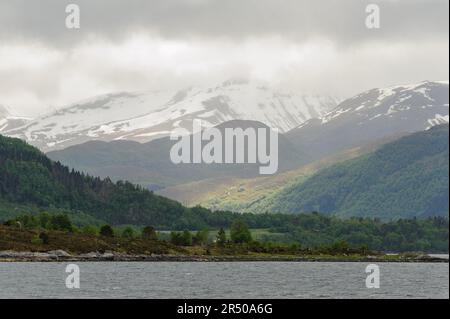 snow-capped mountains with green trees and rays of sunshine break through the clouds Stock Photo