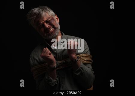 Emotional beaten man tied with rope on black background, space for text ...