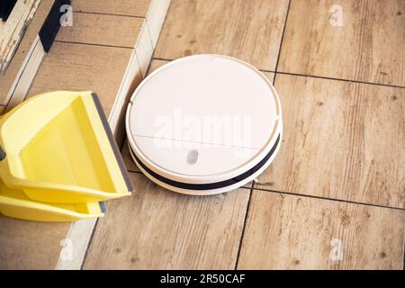 robot vacuum cleaner cleans on a tiled floor in a living room lit by the sun next to yellow dustpans for cleaning, horizontal Stock Photo