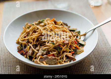 chinese noodles with vegetables and meat on wooden table with natural soft light. stir fry noodles in white dish on table at home. plated and homemade Stock Photo