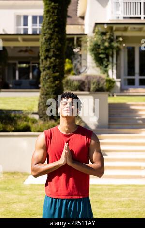 Biracial handsome young man with eyes closed meditating in prayer pose in yard against house Stock Photo