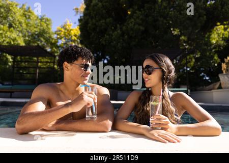 Biracial young couple wearing sunglasses with lemonade talking while standing in swimming pool Stock Photo