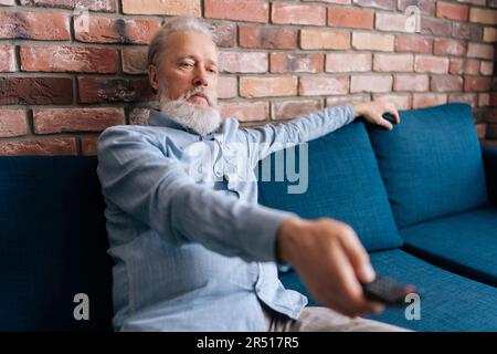Medium shot of bearded mature adult male sitting on couch watching TV enjoying relaxing at home. Gray-haired senior man using remote control to switch Stock Photo