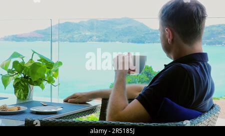 A young man on vacation sits in hotel cafe terrace with view of sea Stock Photo
