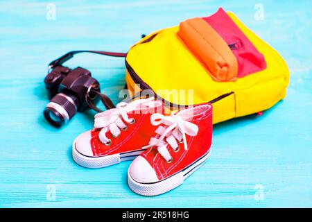 Pair of miniature red shoes, a small colorful backpack, and a toy camera arranged on a blue wooden background. Perfect backdrop for back-to-school or Stock Photo