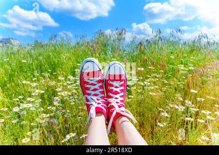 Pair of feet, adorned in red canvas shoes, lay amidst a vibrant field of daisies, with a picturesque view of the blue sky. Stock Photo