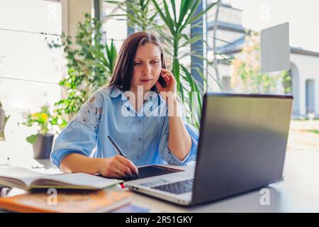 Young female teacher getting ready for lesson working in office using laptop computer. Woman writing notes on tablet Stock Photo