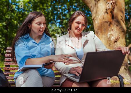 Two young businesswomen cowokers working in spring park using laptop and book. Remote work during break. Friends discussing tasks Stock Photo