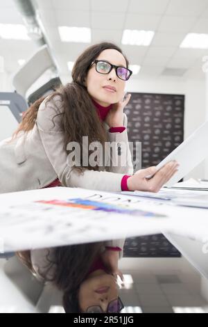 Thoughtful confident beautiful mixed race designer in eyeglasses using tablet and leaning on table with banner in printing house Stock Photo