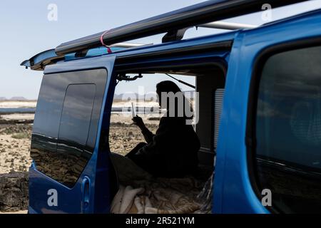 Unrecognizable silhouette of female traveler messaging on smartphone while sitting in blue camper van parked on sunny terrain during summer trip Stock Photo