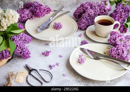 Front view of elegant table setting with fresh lilac flowers, plates with cutlery, scissors and cup of tea on concrete background Stock Photo