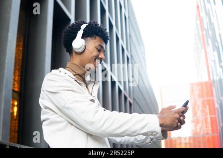 Low angle of positive young African-American man with wireless headphones and afro hairstyle listening to music and smiling while taking a selfie Stock Photo