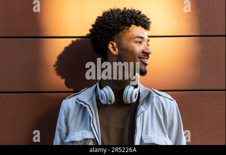 Happy African American young man with afro hairdo and headphones around his neck smiling and looking away from a wall on a street in the sunlight Stock Photo
