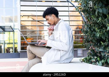 Side view of young African American freelancer with curly dark hair sitting on a bench and browsing his laptop computer Stock Photo