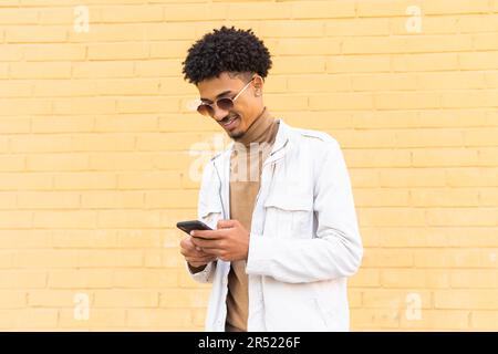 Positive African American young man with sunglasses combed afro sending messages on his mobile phone while standing in the street against a wall Stock Photo