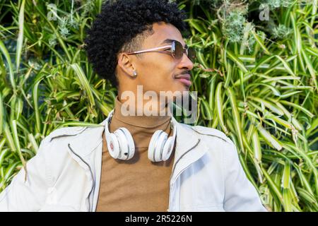 Young African American man in sunglasses with headphones around his neck and curly hair looking away thoughtfully while sitting on the street Stock Photo