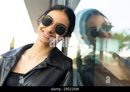 Smiling female with black hair wearing sunglasses and leather jacket standing near window on street Stock Photo