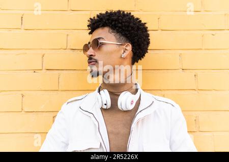 Serious young African American man with sunglasses afro hairstyle and headphones leaning against a brick wall in a sunny street Stock Photo