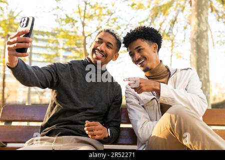 Young African American friends smiling and taking a selfie with their mobile phone sitting on a wooden bench in a sunny park Stock Photo