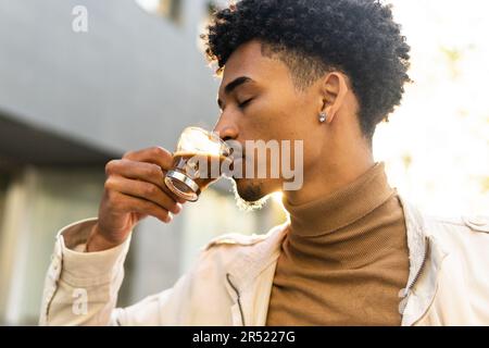From below serious young African American man with afro hairstyle drinking hot coffee in a clear cup with his eyes closed on a sunny street Stock Photo