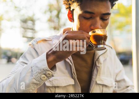 Young African American man with afro hairstyle drinking hot coffee in a clear cup with his eyes closed on a sunny street Stock Photo