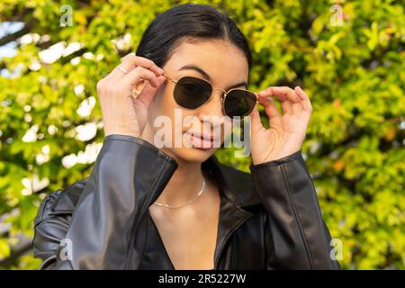 Portrait of confident young female with black hair in leather jacket putting on sunglasses while standing near green bushes Stock Photo