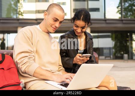 Positive young boyfriend in beige costume and girlfriend in leather jacket sitting on bench near modern building and watching video on laptop Stock Photo