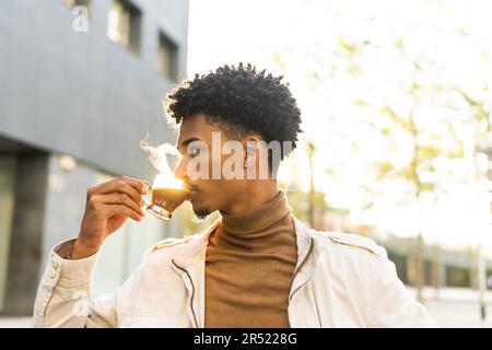 Thoughtful young African American male in casual wear with Afro hairstyle drinking hot coffee from transparent cup while looking away on sunny street Stock Photo
