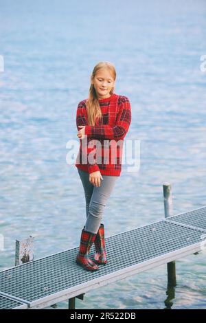 Outdoor portrait of cute little girl posing next to lake on dock, wearing red sweater and rain boots, cold fall or spring weather Stock Photo