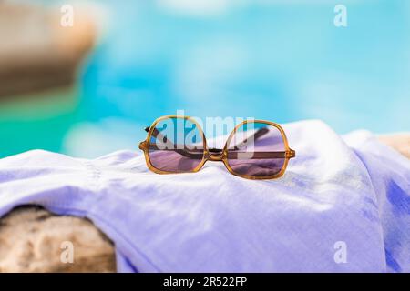 Close up of fashionable hexagonal purple sunglasses with folded arms placed on a white towel on a stone against blue water faded in the sunlight Stock Photo