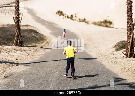 Back view of unrecognizable boy and girl in casual clothes riding kick scooters on empty road while having fun together during sunny day Stock Photo