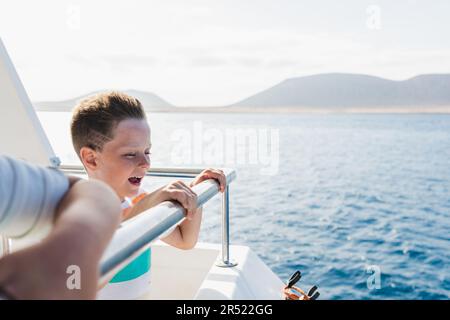 Side view of happy boy in casual clothes smiling and enjoying fresh air while standing on ship deck floating sea water during sunny day Stock Photo