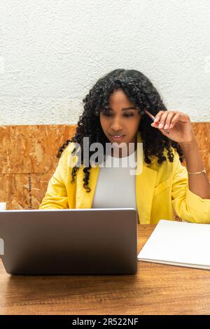 Thoughtful young African American female manager in sitting at wooden table with pen and browsing netbook while working on business startup strategy Stock Photo