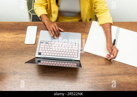 High angle of anonymous African American female sitting at wooden table with smartphone and writing information in copybook while working on laptop Stock Photo