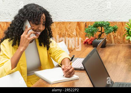 Happy African American young freelancer in smiling and writing information in a diary while talking on a smartphone at a table with two laptops Stock Photo