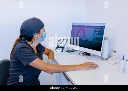 Side view of female dentist in medical mask sitting on chair while using computer and working in bright modern clinic Stock Photo