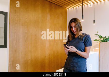 Positive young female medic in uniform smiling and browsing smartphone while standing near counter in modern clinic during break from work Stock Photo