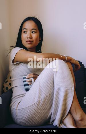 Side view of young pensive confident Asian female in casual clothes with legs up on sofa while looking away in living room indoors Stock Photo