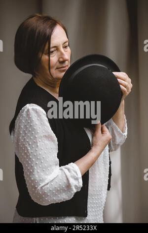 Thoughtful middle aged female model in elegant outfit with black hat and short brown hair looking away against beige curtain in studio Stock Photo